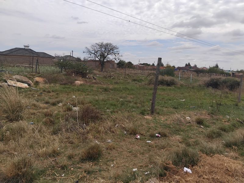 A vacant residential plot featuring natural grass, scattered debris, and basic wire fencing with overhead power lines visible.