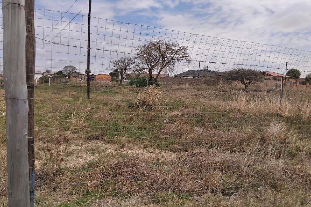 A view of a vacant residential plot enclosed by wire mesh fencing and wooden posts, featuring natural grass and scattered trees.