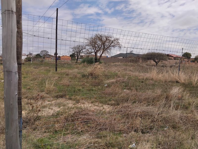 A view of a vacant residential plot enclosed by wire mesh fencing and wooden posts, featuring natural grass and scattered trees.