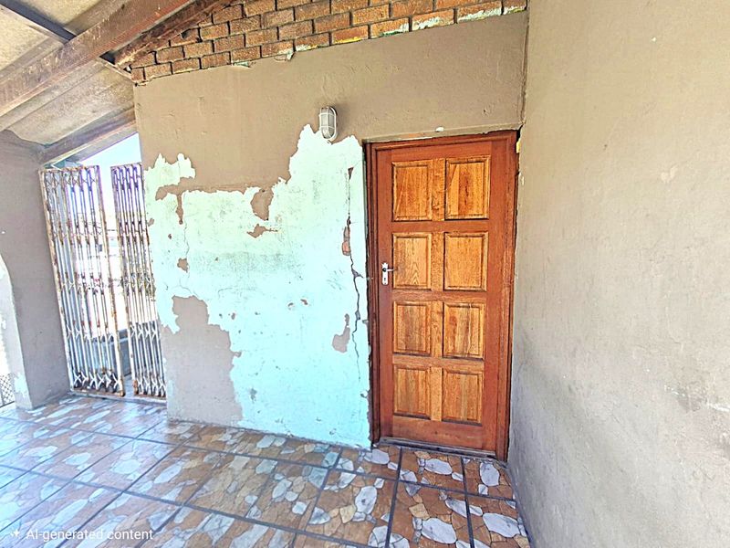 A covered patio area featuring tiled flooring, a wooden entrance door, and a security gate, currently requiring maintenance to the wall finishes.