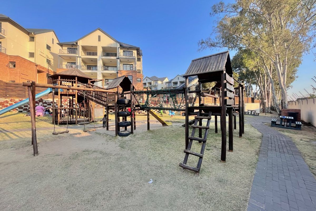 Communal outdoor area featuring a wooden playground structure, paved pathways, and landscaped grounds within an apartment complex setting.