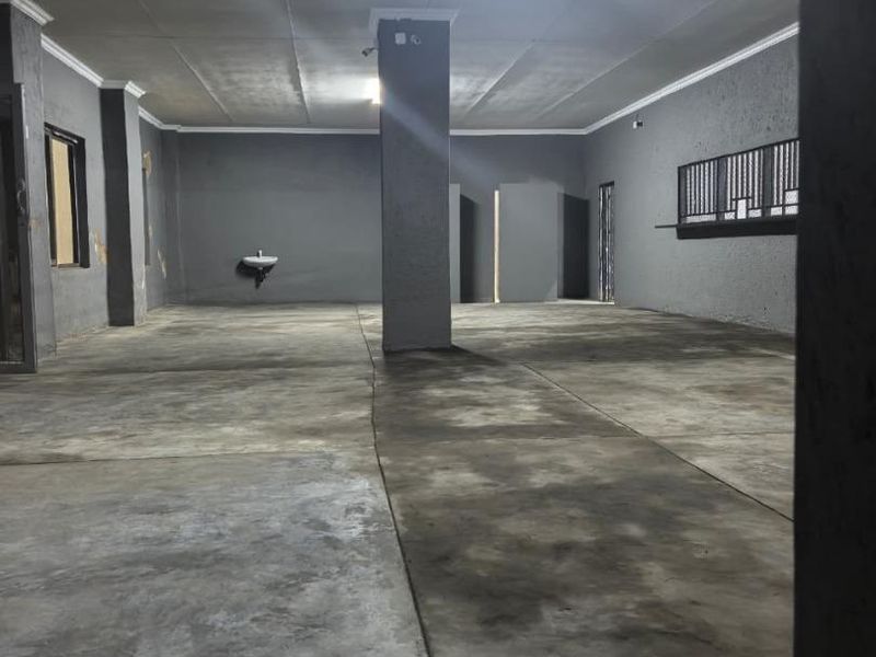 A spacious, empty commercial open-plan area featuring concrete flooring, a wall-mounted basin, and a security window grille, suitable for various business applications.
