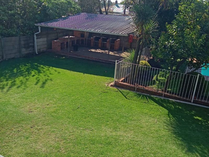 A spacious backyard featuring a well-maintained lawn, mature trees, and a covered patio area with a built-in brick braai.