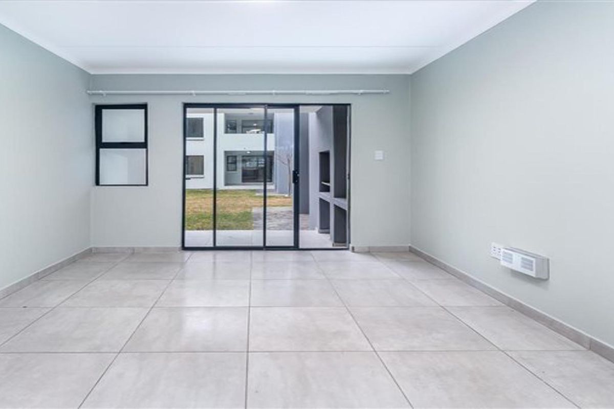 A bright, empty living room featuring neutral tiled floors, a wall-mounted heater, and sliding glass doors leading to an outdoor patio area with a braai.