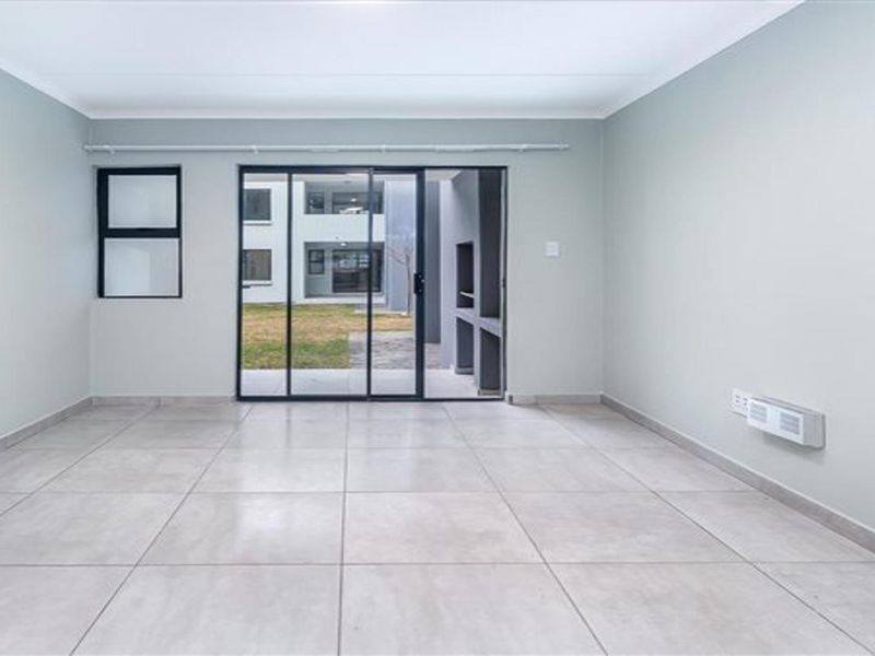 A bright, empty living room featuring neutral tiled floors, a wall-mounted heater, and sliding glass doors leading to an outdoor patio area with a braai.