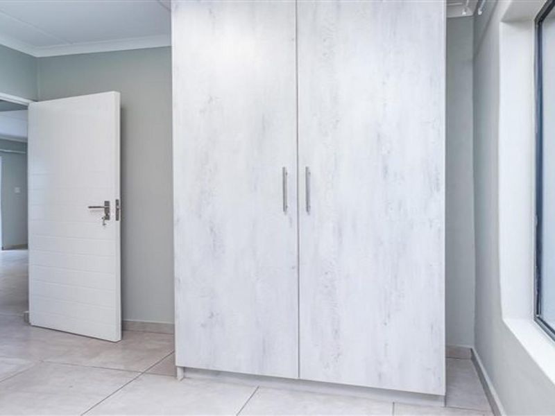 A bright, empty bedroom featuring modern light-wood built-in cupboards, neutral tiled flooring, and large windows allowing for ample natural light.