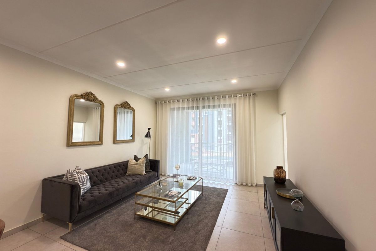 A well-lit living room featuring neutral tiled flooring, recessed ceiling lighting, and a large sliding glass door providing access to the balcony.