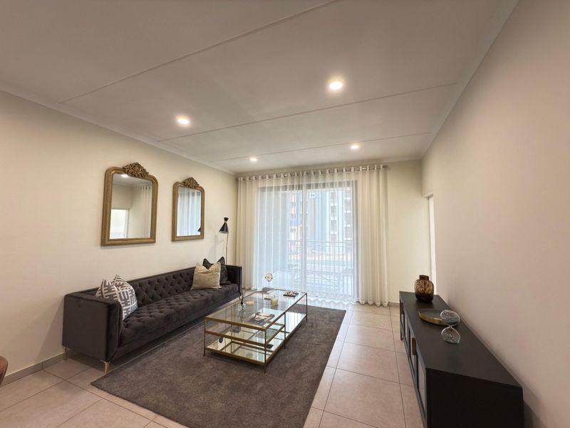 A well-lit living room featuring neutral tiled flooring, recessed ceiling lighting, and a large sliding glass door providing access to the balcony.
