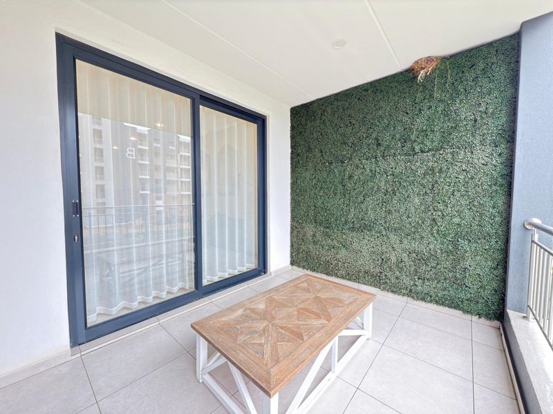 A covered balcony featuring tiled flooring, a decorative artificial green wall, and a sliding glass door providing access to the apartment interior.