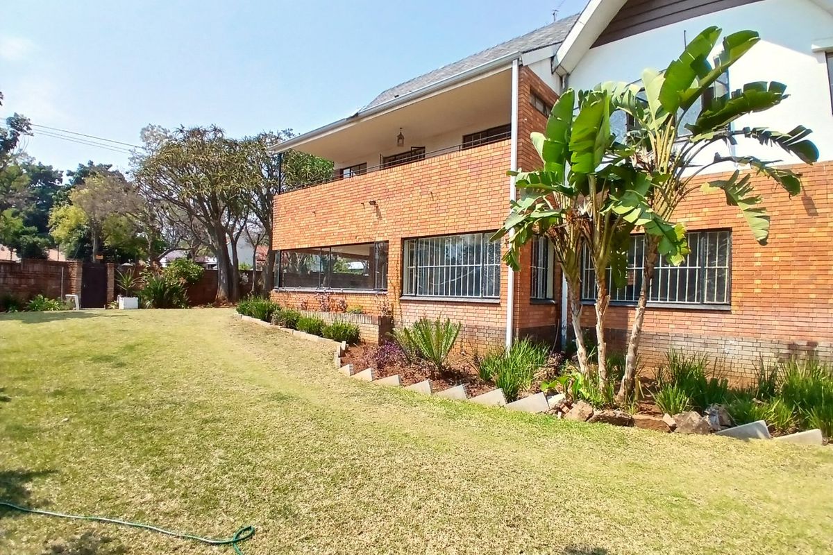 Rear view of a double-story brick house featuring a spacious lawn, landscaped garden beds, and an upper-level balcony with security bars on windows.