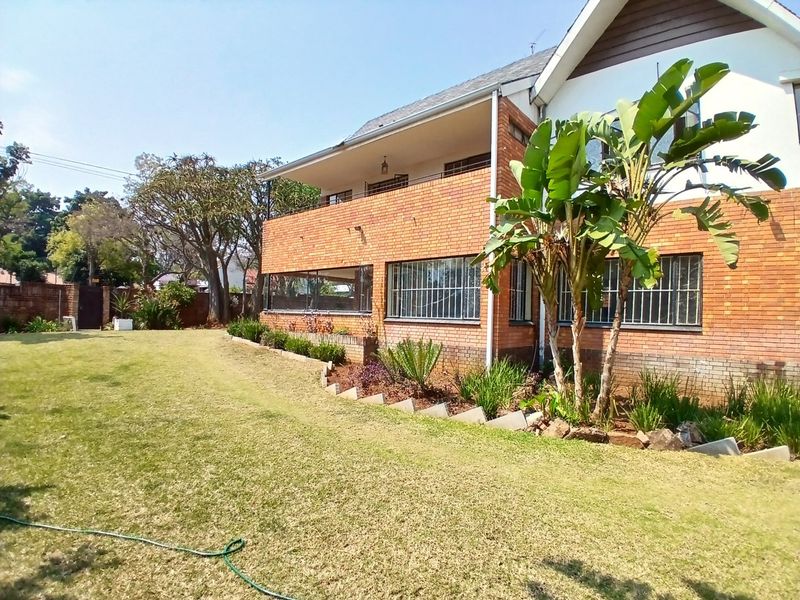 Rear view of a double-story brick house featuring a spacious lawn, landscaped garden beds, and an upper-level balcony with security bars on windows.