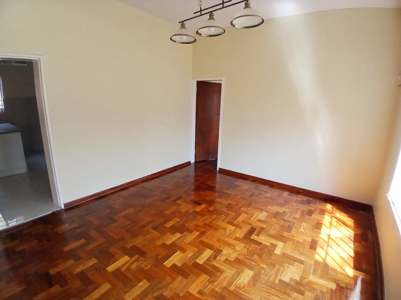 A bright, empty living room featuring polished parquet timber flooring, neutral walls, and a classic pendant light fixture.