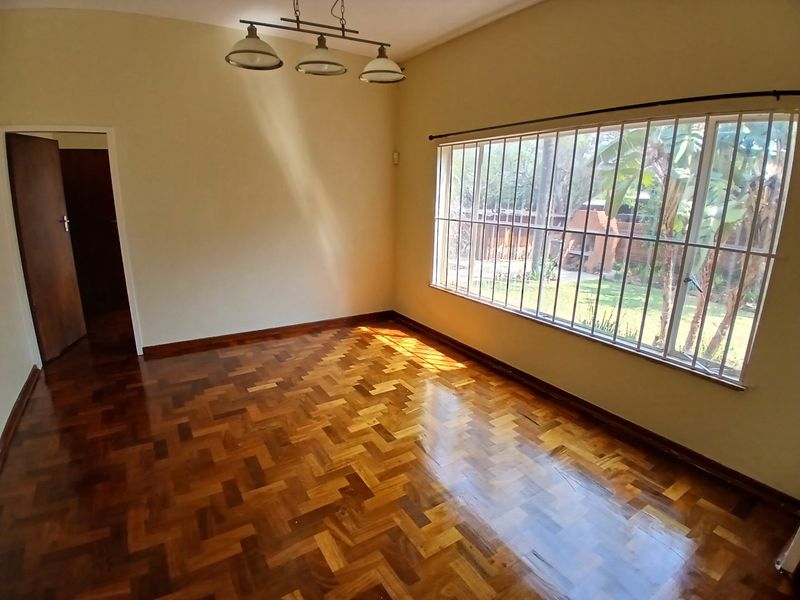 A bright, empty living room featuring polished parquet flooring, a large window with security bars, and a central pendant light fixture.