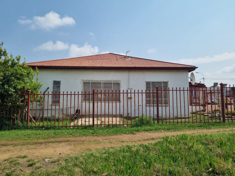 Front view of a single-story house featuring a corrugated metal roof, security bars on windows, and a metal perimeter fence.