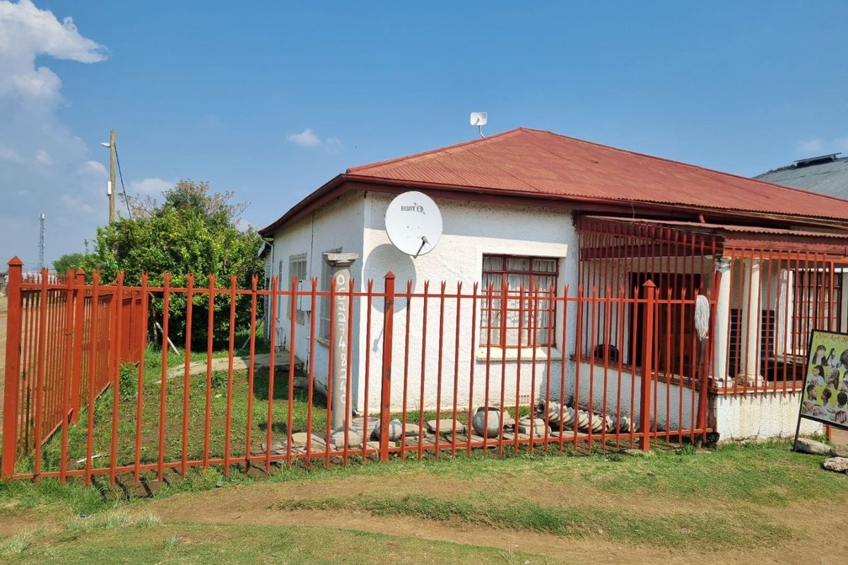 Front view of a single-story house featuring a corrugated metal roof, security fencing, and a concrete pillar entrance.