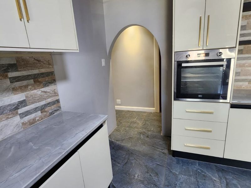 A modern kitchen featuring built-in cabinetry, a wall-mounted oven, stone-look countertops, tiled backsplash, and a distinctive timber ceiling with an arched entryway.