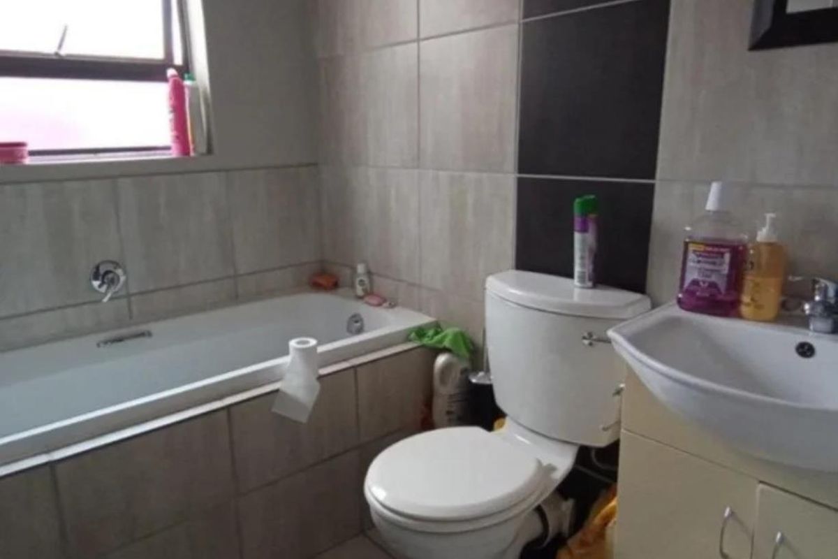 A functional bathroom featuring a bathtub, toilet, and vanity basin with neutral wall tiling and a window for natural light.