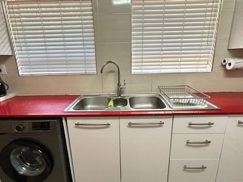 A functional kitchen area featuring white cabinetry, a striking red quartz countertop, double stainless steel sink, and integrated washing machine with dual window access.