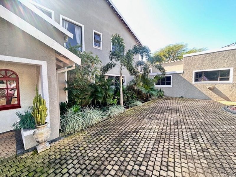 Front exterior view of the house featuring a cobblestone driveway, landscaped garden beds, and a stucco facade with arched and casement windows.