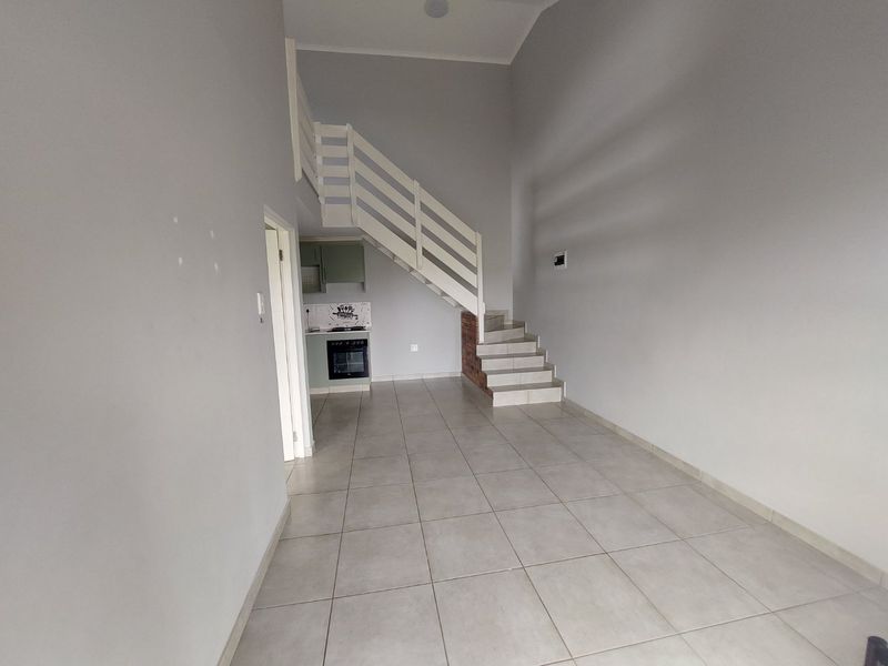 An open-plan living area featuring tiled flooring, a staircase leading to an upper level, and a compact kitchen with built-in appliances.