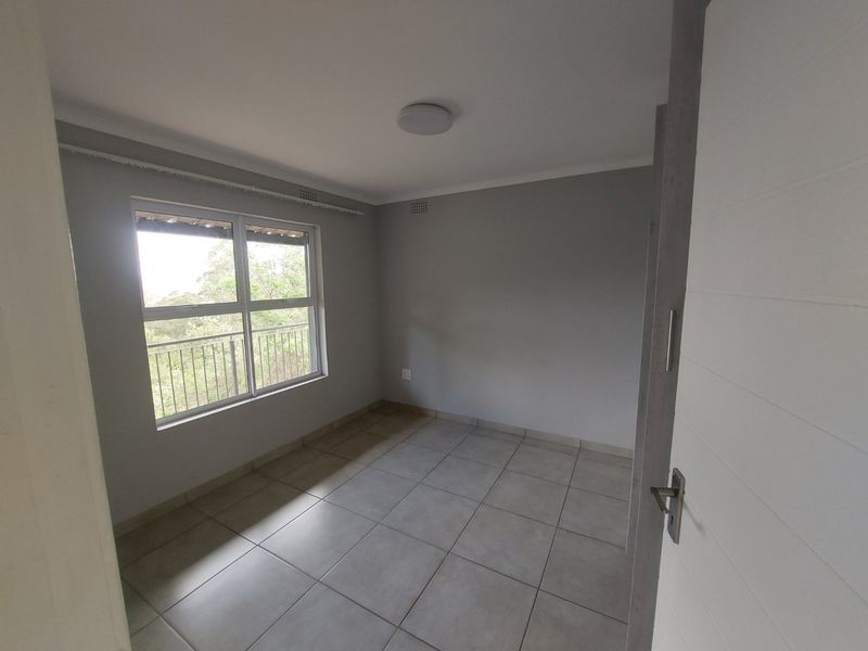 A clean, empty bedroom featuring neutral grey walls, tiled flooring, and a large window with security bars overlooking greenery.