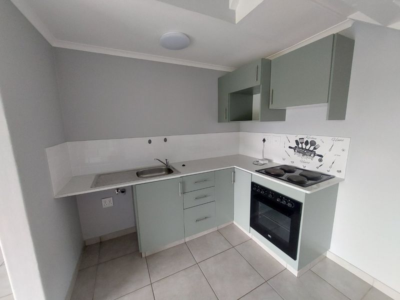 A compact, modern kitchen featuring sage green cabinetry, a built-in oven and hob, stainless steel sink, and neutral tiled flooring.