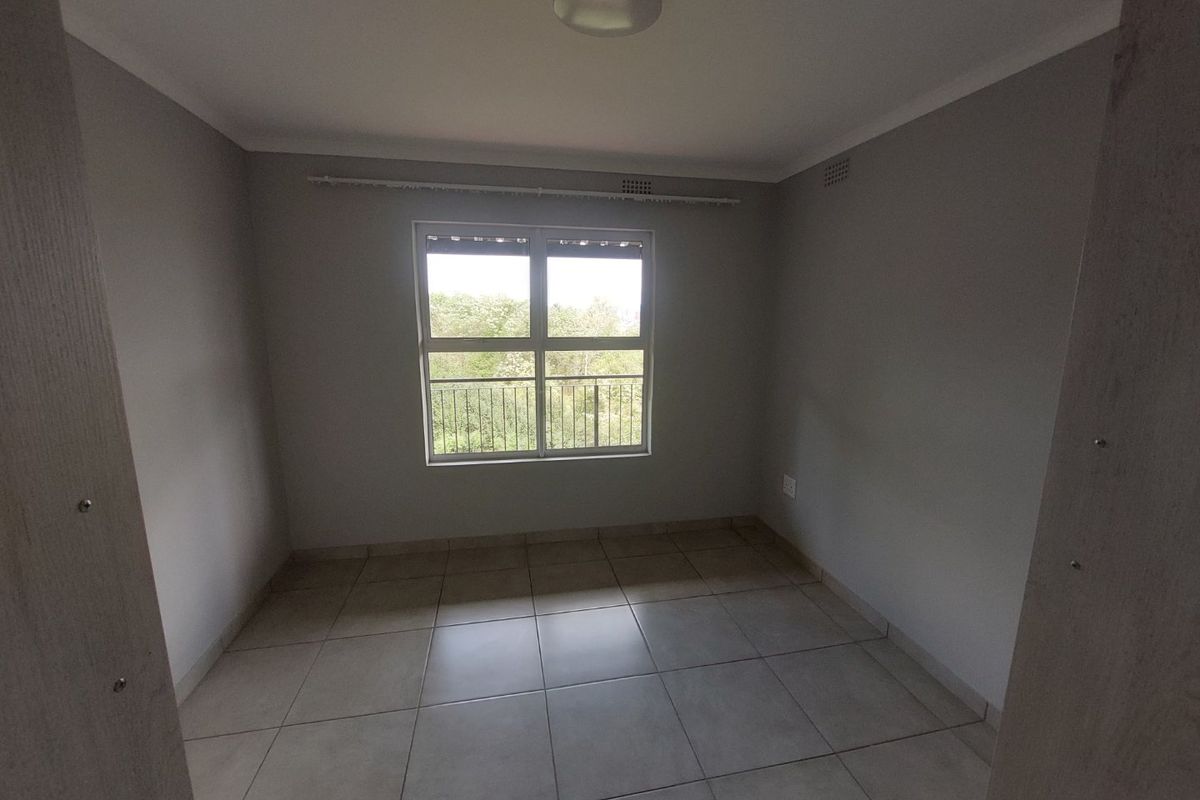 A clean, empty bedroom featuring neutral tiled flooring, grey walls, and a large window providing natural light and an exterior view.