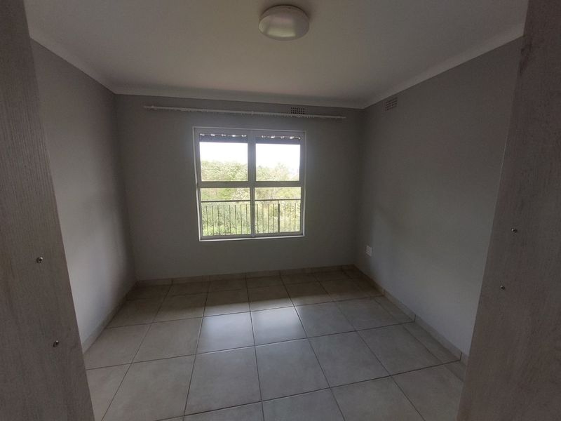 A clean, empty bedroom featuring neutral tiled flooring, grey walls, and a large window providing natural light and an exterior view.