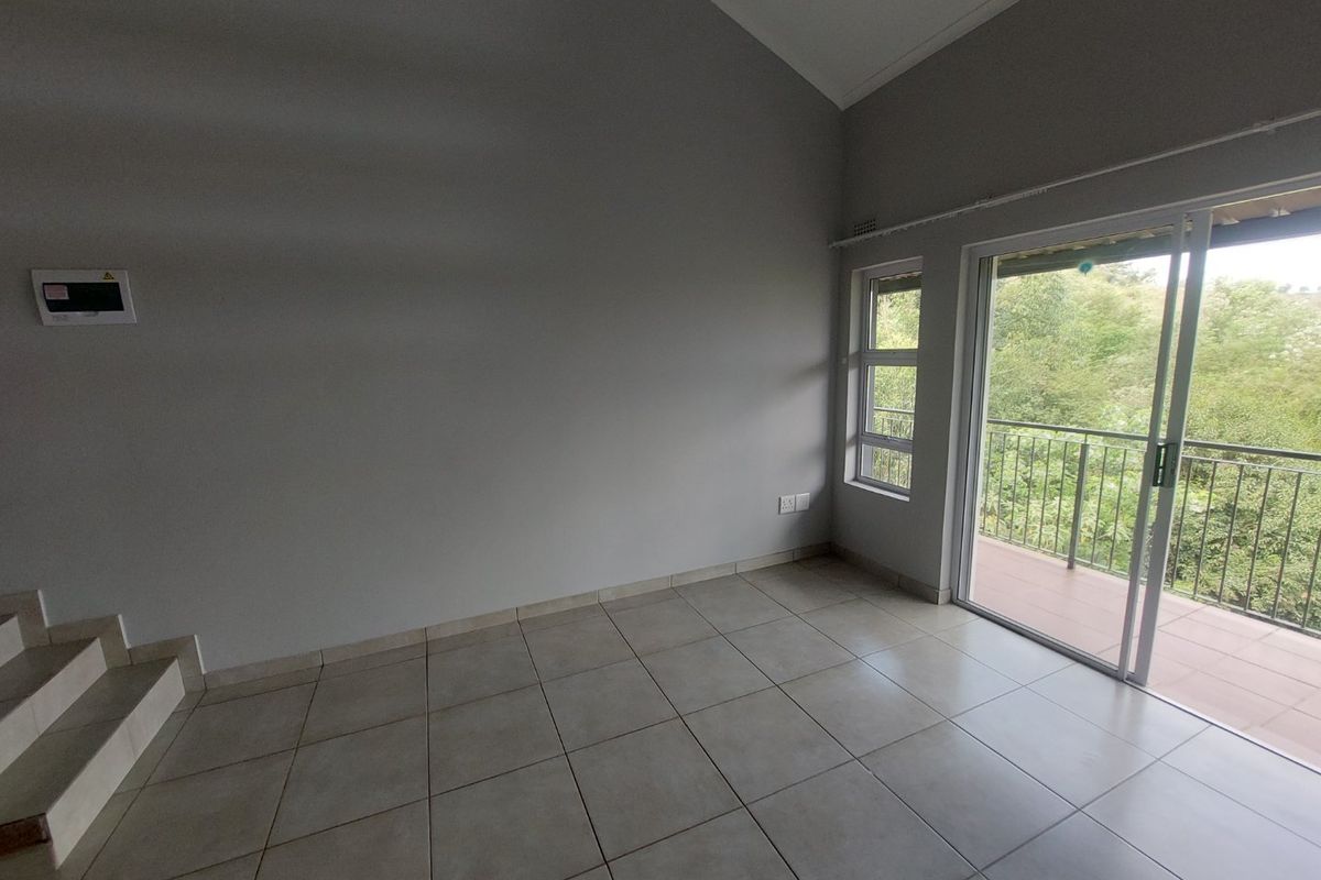An empty living room featuring tiled flooring, a vaulted ceiling, and a sliding glass door providing access to a balcony with greenery views.