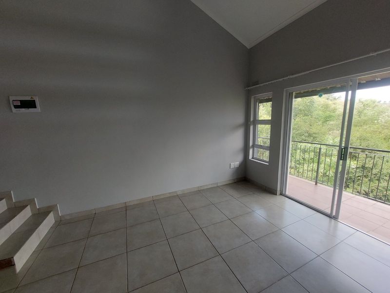 An empty living room featuring tiled flooring, a vaulted ceiling, and a sliding glass door providing access to a balcony with greenery views.