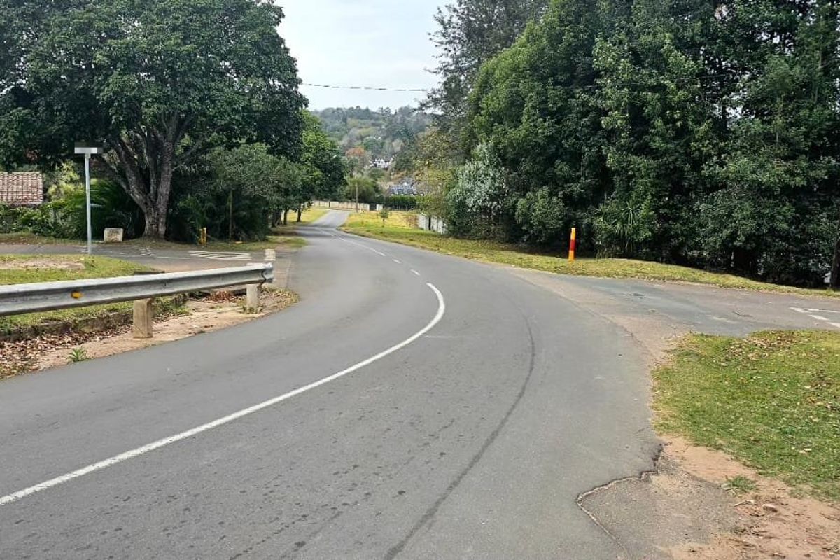 A view of the local street approach leading towards the property, featuring asphalt paving, roadside guardrails, and surrounding mature trees.