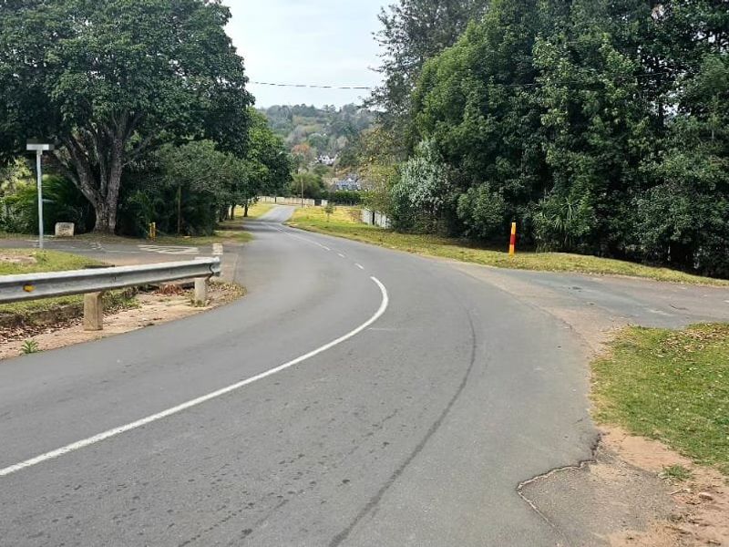 A view of the local street approach leading towards the property, featuring asphalt paving, roadside guardrails, and surrounding mature trees.