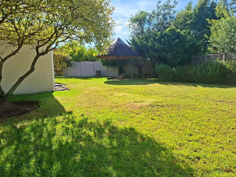 A spacious private garden featuring a well-maintained lawn, mature trees, and a traditional thatched lapa area for outdoor relaxation.