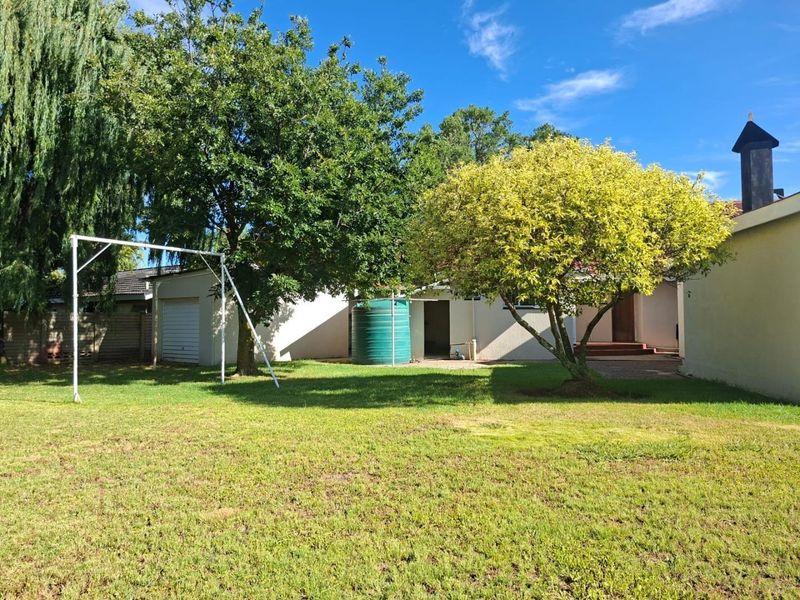 A spacious rear garden featuring a lawn, mature trees, a water tank, and access to the garage and main house structure.