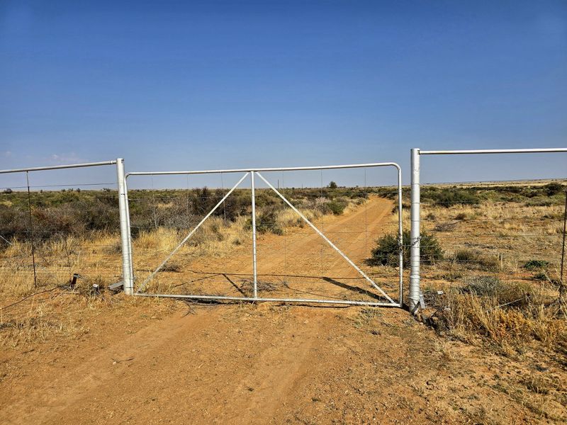 A functional metal farm gate providing access to the property via a dirt track, set within standard wire fencing in a rural landscape.