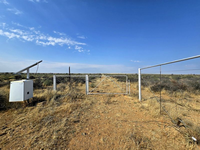 A rural farm entrance featuring a metal gate, wire fencing, and a solar-powered electrical control unit set against an open landscape.