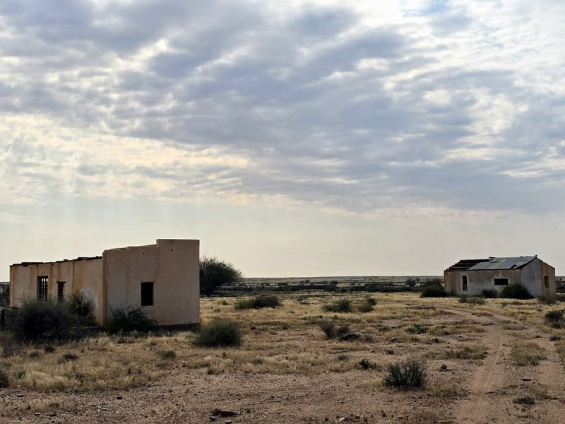 A view of two dilapidated farm structures set in a vast, arid landscape, showing significant structural deterioration and weathered exterior finishes.