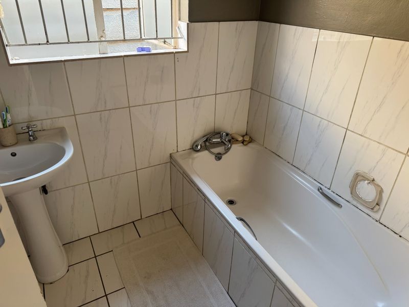 A functional bathroom featuring a pedestal basin, bathtub with chrome fixtures, and marble-effect wall tiling with security bars on the window.