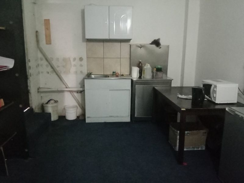 A basic kitchenette area featuring a sink, wall-mounted cabinets, and a stainless steel prep station, requiring cosmetic updates and wall repairs.
