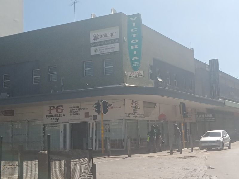 Street-level view of a commercial corner building featuring prominent signage, security shutters, and an awning, located in a busy urban area.