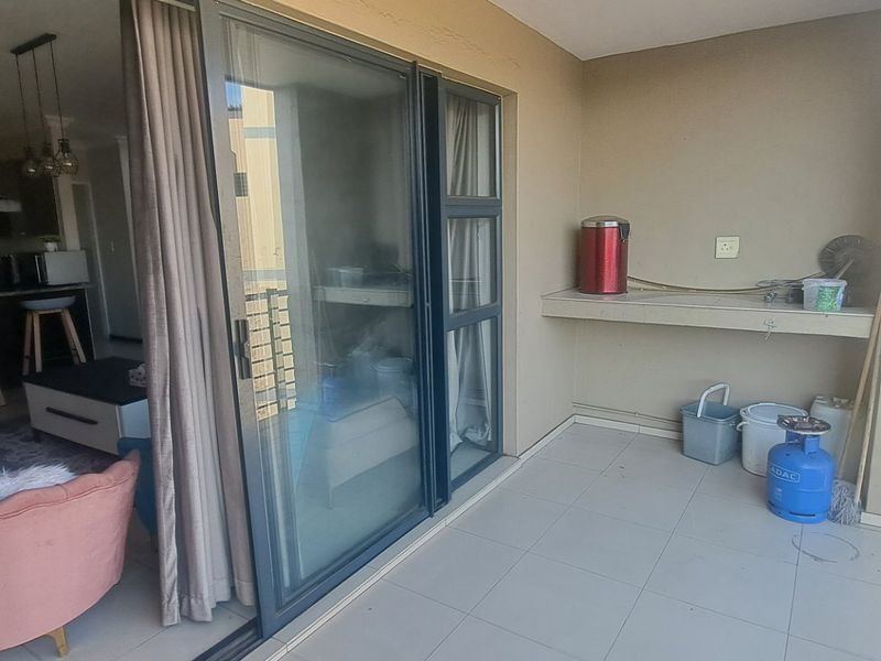 A covered balcony featuring tiled flooring, a built-in counter, and a metal railing, providing seamless access to the adjacent open-plan living and kitchen area.
