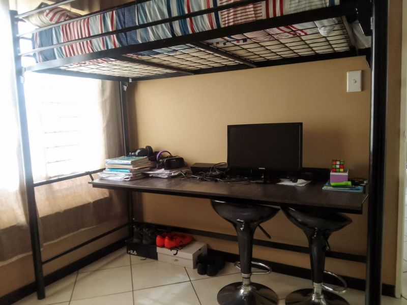 A bedroom featuring tiled flooring and a louvered window, currently utilized with a loft bed and integrated desk workspace.