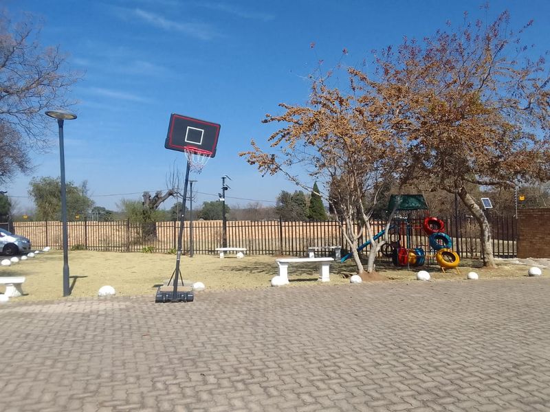 A communal outdoor area featuring a paved surface, perimeter fencing, concrete seating, and recreational equipment within a residential townhouse complex.
