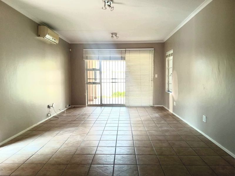 An empty, neutrally painted living room featuring tiled flooring, a wall-mounted air conditioning unit, and a sliding glass door with security gate access.