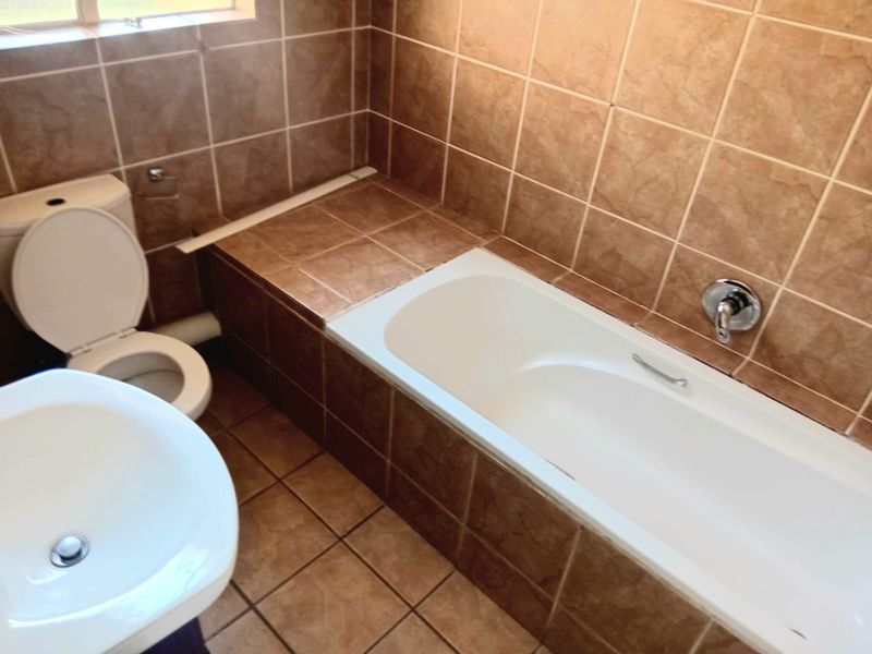 A functional bathroom featuring a built-in bathtub, pedestal basin, and toilet, finished with brown ceramic wall and floor tiles.