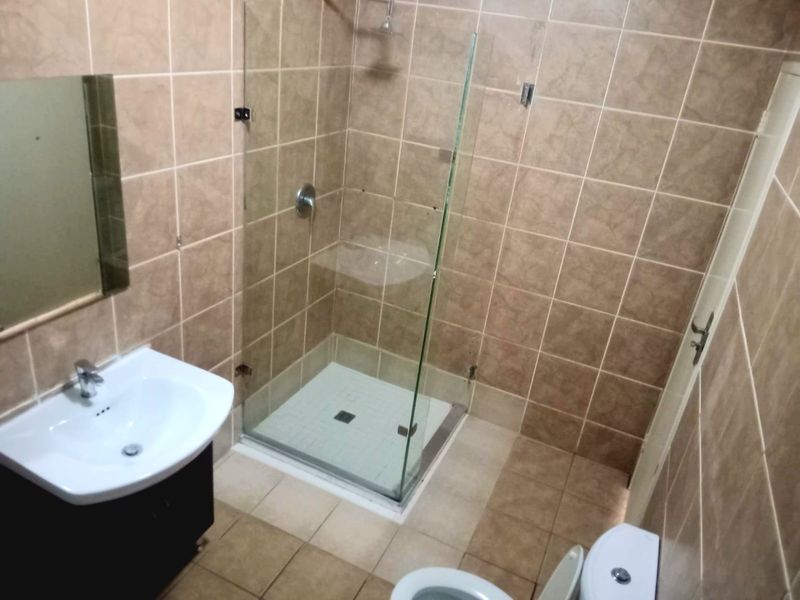 A functional bathroom featuring a glass-enclosed shower, vanity basin, and toilet with neutral wall and floor tiling.