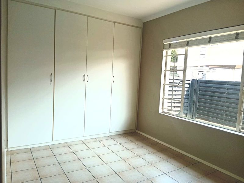 A bright, empty bedroom featuring neutral walls, tiled flooring, and ample built-in cupboard storage with security bars on the window.