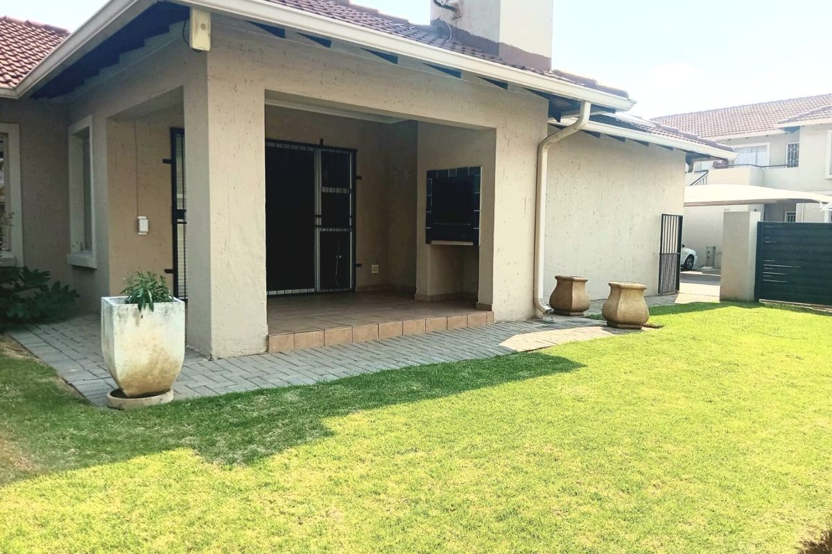 Rear view of the townhouse featuring a covered patio with a built-in braai, paved walkway, and a well-maintained garden area.