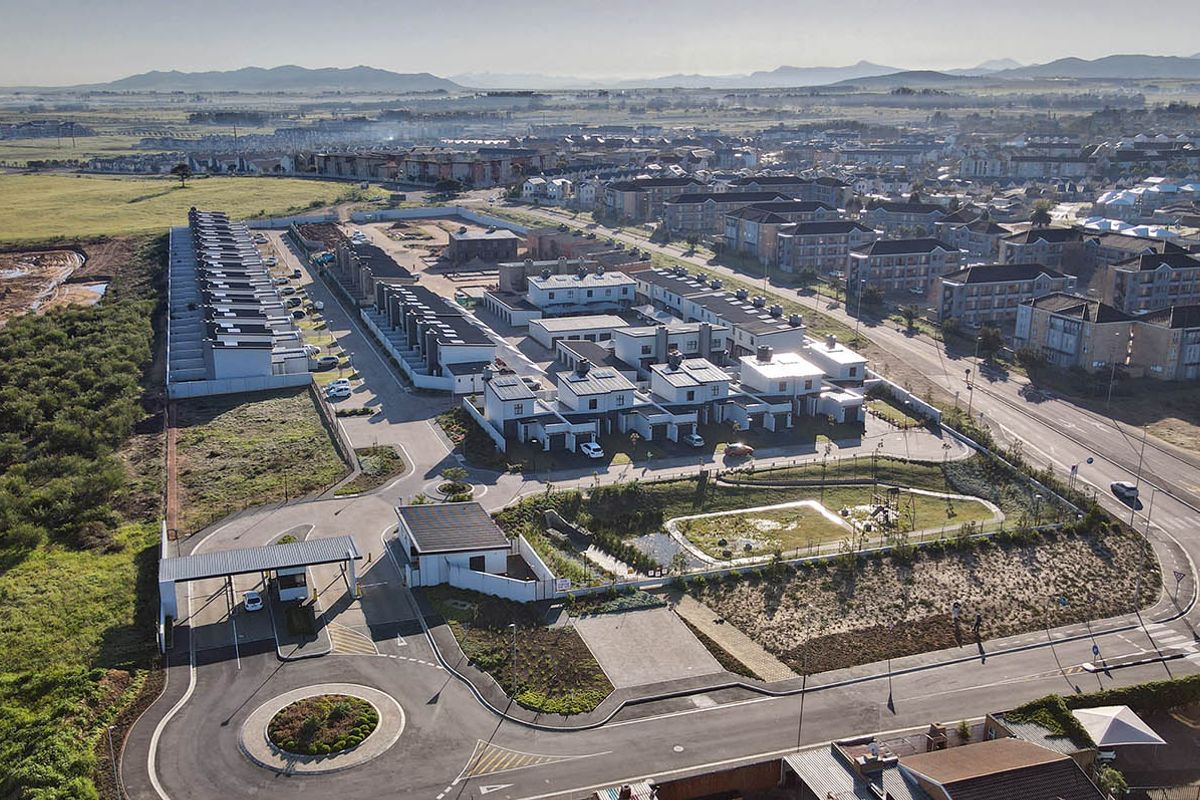 Aerial view of a modern residential townhouse complex featuring a secure gated entrance, paved internal roads, and landscaped communal green spaces.