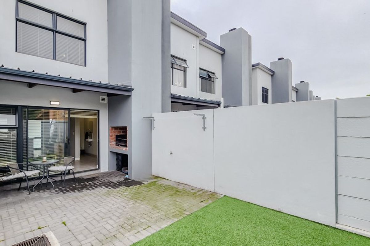 Rear townhouse exterior featuring a paved patio area with a built-in braai, artificial lawn, and sliding door access to the interior living space.
